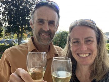 A woman and a man are smiling at the camera and holding up glasses of champagne. They are outside in the sun. Both people have sunglasses on their heads.
