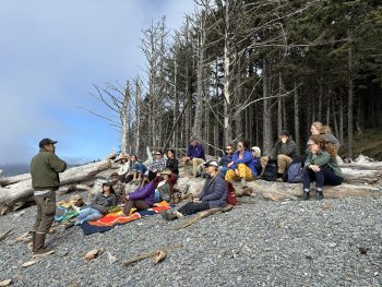 The fellows sit on a beach with a forest in the background