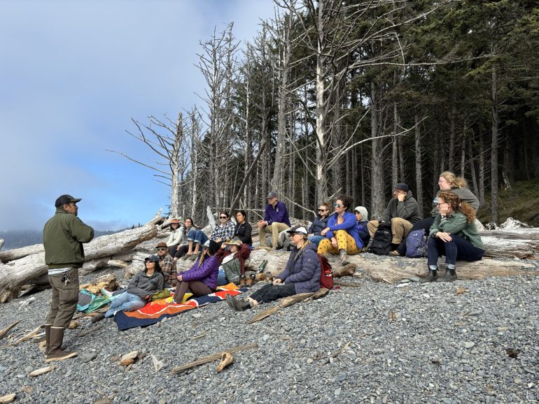The fellows sit on a beach with a forest in the background