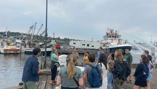 The cohort stands on a dock with a boat in the background
